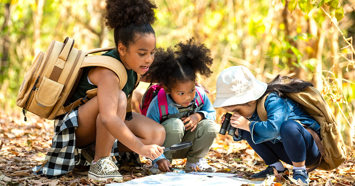 Family Fitness_blog-4 Children with backpacks explore a wooded trail, using a magnifying glass and binoculars to examine nature.