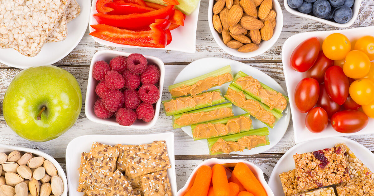 Assortment of healthy snacks including fresh fruits, vegetables, nuts, crackers, and celery topped with peanut butter.