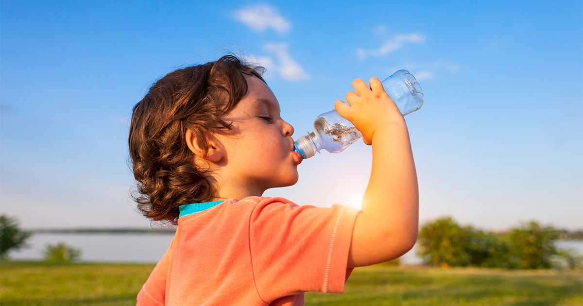 Child drinking water from a plastic bottle outdoors on a sunny day.